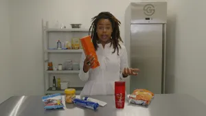 Een foto van Eva die in een witte jurk achter een stalen tafel staat. Ze heeft een pak crackers vast. Op de tafel liggen zes verpakte producten waarin, net als in de crackers, palmolie zit: mayonaise, chips, koekjes, shampoo, snoepjes en pindakaas. 