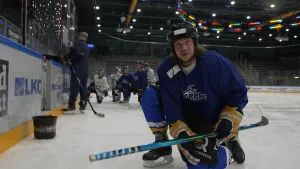 Een foto van Pascal die op zijn knie op een ijshockeybaan zit in een blauw pak met schaatsen, grote handschoenen en een helm. Hij heeft een ijshockeystick vast om de puck, een ronde schijf, te slaan. Op de achtergrond krijgen spelers uitleg van de coach.