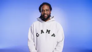 Een foto van de Nederlandse rapper en zanger Glen Faria die lacht naar de camera. Hij heeft een witte capuchontrui aan met in het zwart de letters "CAMP" erop. De achtergrond van de foto is lichtblauw.