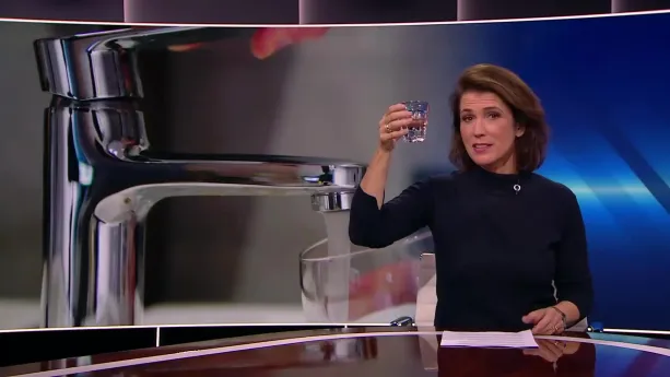 Een foto van de presentator die aan tafel zit in de studio. Ze houdt een glas met water omhoog met haar rechterhand. Op de achtergrond is er een open keukenkraan te zien waar een glas onder wordt gehouden.