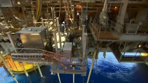 Een foto van een boortoren waar aardolie gewonnen wordt. Er zijn drie medewerkers te zien op de foto die aan het werk zijn en helmen op hebben. Onderaan de foto is zeewater te zien.