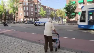 Een foto van een oude mevrouw die achter een rollator, een looprek met wielen, loopt op de stoep. De mevrouw wil oversteken. De wielen van de rollator raken bijna een rood fietspad waarachter een tram en auto naar links rijden en een scooter naar rechts.