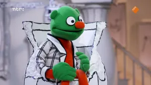 Een foto van een pop in een witte stoel. De pop heeft een groene huid, een kaal hoofd, grote, ronde ogen en een donkerrode neus. Hij draagt een wit jasje met zwarte lijnen erop die door elkaar heen lopen en rechthoeken vormen.