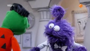 Een foto van twee poppen. De pop links heeft een groene huid, zwart stekelhaar en een rode trui met zwarte driehoeken erop. De pop kijkt naar de pop rechts met een paarse vacht, een krul op zijn hoofd, een witte strik om zijn hals en een wit jasje aan.