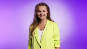 Een foto van zangeres en actrice Vajèn van den Bosch. Ze lacht naar de camera en heeft een groengele blouse aan. Ze staat voor een paarse achtergrond.