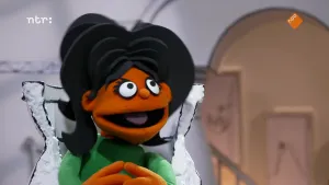 Een foto van een pop in een witte stoel. De pop heeft hoog zwart haar, met zwarte lokken naast het hoofd. De pop draagt een groene trui, heeft een bruine huid, ronde ogen met wimpers, een kleine, ronde neus en een open mond met een roze tong erin.