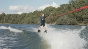 Een foto van presentator Pascal die aan het waterskiën is. Hij houdt met een hand de lijn vast terwijl hij zijn andere hand in de lucht houdt.