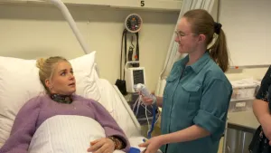 Een foto van presentator Janouk die in een ziekenhuis bed ligt. Naast haar staat een student die een apparaat in haar hand heeft waarmee je in het oor de temperatuur van het lichaam kan opmeten.