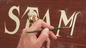 Een foto van een hand die op een bruine houten plank witte letters met een dunne kwast schildert en ook een stok met een lapje stof vastheeft om de plank niet te raken. Letter "M" wordt op de foto geschilderd, samen met letters ervoor is "Stam" te lezen.