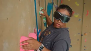 Een foto van de presentator die met een skibril op waar je niet doorheen kan kijken aan het klimmen is aan een klimmuur.