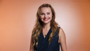 Een foto van zangeres en actrice Vajèn van den Bosch. Ze lacht naar de camera en heeft een donker vest aan. Ze staat voor een oranjebruine achtergrond.