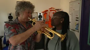 Een foto van presentator Tirsa die binnen naast een man staat die haar laat zien hoe je trompet moet spelen.