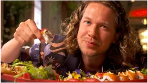 Een foto van presentator Pascal die dicht op de camera zit met een stuk Sushi in zijn hand. Voor hem ligt er nog meer Sushi en groente. Sushi is een Japanse maaltijd dat bestaat uit rauwe vis.