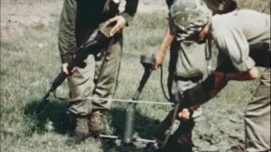 Een foto van soldaten die naar mijnen zoeken. Een van de soldaten houdt een gereedschap tegen de grond waarmee naar mijnen wordt gezocht.