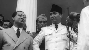 Een foto van president Soekarno die voor een microfoon staat. Naast hem en achter hem staan er nog meer mannen.