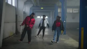 Een foto van een presentator tussen twee dansers in een leeg magazijn. Ze zijn een danspas aan het uitvoeren waarbij ze de linkervoet op de grond hebben en de rechtervoet met de hak op de grond zetten. Op de achtergrond komt licht door een garagedeur.