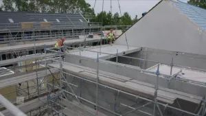 Een foto van een bouwterrein waar huizen gebouwd worden. Er zijn twee bouw medewerkers te zien die bezig zijn met een grote betonnen plaat die aan een kraan hangt.