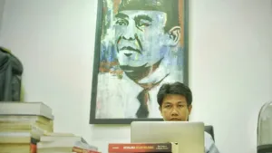 Een foto van een man die zit op een bureaustoel achter een laptop met voor zich drie stapels boeken. Op de achtergrond hangt een geschilderd portret van Soekarno. Hij heeft op het schilderij een zwarte hoed op, een witte blouse aan en een stropdas om.