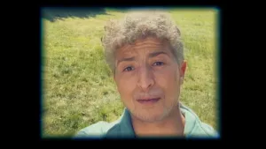 Een foto van een oudere man met grijs haar die vlak voor de camera op een grasveld staat.