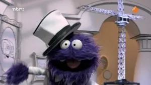 Een foto van een pop met een harige, paarse vacht en een witte hoed op. De pop heeft zijn mond open en zijn twee boventanden zijn te zien. Op de achtergrond staat rechts een grote, witte bouwkraan. Links hangt een schilderijlijst met een caravan erop.