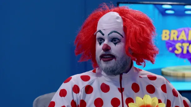 Een foto van een clown met een rode pruik op zijn hoofd die een wit vest met rode bolletjes erop aanheeft. De clown heeft een wit beschilderd gezicht, waarop de neus en lippen rood geschilderd zijn en de wenkbrauwen en de randen om de ogen zwart. 