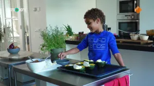 Een foto van een jongetje dat kookt in de keuken en een beetje kruiden tussen twee vingers heeft. Voor hem staan, naast tijm en uien, zes met kaas en kruiden opgevulde helften kleine, groene pompoentjes op een bakplaat voor in de oven. 