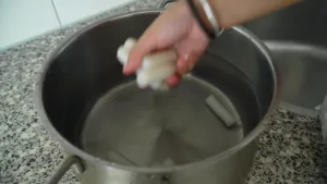 Een foto van een stalen pan op een aanrecht. In de pan zit een laagje water en liggen een aantal witte rijstcake sticks, dat is rijst in de vorm van een staafje. Een hand van het meisje dat kookt houdt vier rijstcake sticks vast boven de pan.