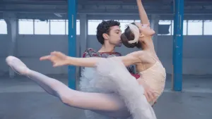 Een foto van een man en een vrouw die in een lege magazijnhal klassiek ballet aan het dansen zijn in balletkleding. De vrouw staat in een positie met het rechterbeen en de rechterarm naar achteren gestrekt en de linkerarm en het hoofd omhooggeheven. 