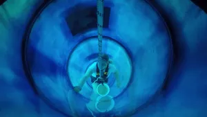 Een foto van een vrouw die aan een dik touw hangt in het midden van de blauwe buis van een waterglijbaan. De vrouw is de waterglijbaan aan het schoonmaken met een spons. Aan het touw hangt ook een emmer met schoonmaakmiddel erin.