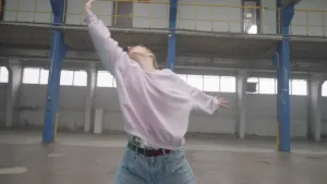 Een foto van een danseres die in een lege magazijnhal urban contemporary, een mix van verschillende dansstijlen, danst. Ze kijkt met haar schuin omhoog geheven hoofd naar haar rechts geheven rechterhand en strekt de linkerhand links naar achteren.