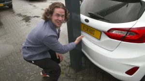 Een foto van presentator Pascal die gehurkt staat en daarbij kijkt naar de camera en met zijn hand wijst naar een witte auto van het merk Ford. De auto staat tegen een grote, zwarte paal aan. Op het gele nummerbord van de auto staat "KLOKHUIS".