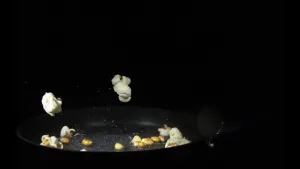 Een foto van een zwarte koekenpan met popcorn erin. Er liggen losse maïsstukjes in uit het binnenste van de popcorn en ook stukjes hele popcorn. Op de foto springen twee stukjes hele popcorn omhoog uit de pan. De achtergrond van de foto is zwart.