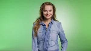 Een foto van zangeres en actrice Vajèn van den Bosch. Ze lacht naar de camera en heeft een blouse van spijkerstof aan. Ze staat voor een groene achtergrond.