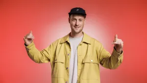 Een foto van muzikant en leraar Willem van der Krabben in een gele blouse met zwarte pet. Hij heeft zijn armen gespreid voor de camera met zijn duim en pink opzij gestrekt en de drie middelste vingers naar voren gebogen in een groet met de naam shaka.