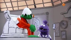 Een foto van twee gekleurde poppen in een zwart-witte kamer. De paarse pop praat tegen de groene pop, die op een hoge stoel zit. Alle dingen in de kamer hebben zwarte lijntjes, alsof ze getekend zijn. 