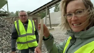 Een foto van een man en een vrouw in een geel hesje met zilveren lijnen erop. De vrouw toont een stukje van de ribben van een Mammoet aan de camera. Het stukje ribben is net door de man gevonden ussen de kiezelstenen die achter hen liggen.