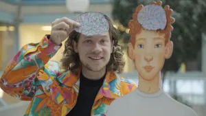 Een foto van Pascal die een getekend stel hersenen voor zijn voorhoofd houdt. Naast hem staat een getekende pop van een jongen met op dezelfde plek ook een stel hersenen. Pascal draagt een blouse met vrolijke kleuren en bladeren erop.