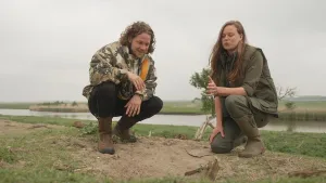 Een foto van presentator Pascal die samen met een boswachter op de grond zit. Op de achtergrond zijn er grasvelden en een rivier te zien.