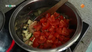 Een foto van een stalen pan op een kookplaat met allemaal ingrediënten erin: kip, aubergine en tomaat zijn te zien. Er zit een houten spatel in de pan. In de pan Een meisje dat niet op de foto staat is pindasoep uit Sierra Leone aan het koken.