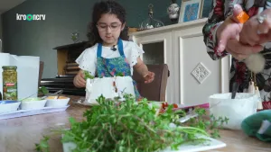 Een foto van een meisje met voor zich op tafel een grote, witte taart met groene blaadjes erop die nog niet helemaal klaar is. Het wordt een Labnehtaart, een taart uit Syrië. Het meisje heeft een blauwe schort met palmbomen erop aan over een witte blouse.