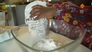 Een foto van een meisje waarvan de rode jurk met bloemen en de linkerhand zichtbaar zijn. Ze heeft een pak met witte bloem vast en ze heeft al een beetje witte bloem in de glazen kom voor zich op tafel gedaan. Ze gaat een Surinaamse pastei maken.