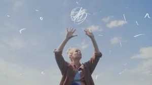 Een foto van Janouk tegen een blauwe lucht met wolken. Janouk houdt haar handen met gespreide vingers omhoog. Er zijn allemaal witte krassen op de foto om Janouk heen te zien om de verschillende broeikasgassen die in de lucht zitten uit te beelden.