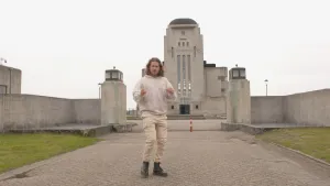 Een foto van presentator Pascal die op een voetpad staat voor Radio Kootwijk. Dat is een heel groot grijs gebouw die in 1918 gebouwd werd.