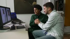 Een foto van presentator Nizar die samen met een man achter een beeldscherm zit terwijl ze elkaars handen vasthouden in een vuist. Op het beeldscherm zijn twee foto's van Nizar te zien. Het beeldscherm is aangesloten op een laptop die ernaast staat.