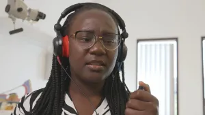 Een foto van presentator Tirsa die ergens binnen staat met een koptelefoon op. Ze houdt een schakelaar vast en heeft een duim op de knop.