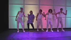 Een foto van Pascal die met vier dansers van de dansgroep 155 danst. Allemaal houden ze één vuist omhoog. Ze staan voor een achtergrond met oplichtende kleuren in vierkante vormen. De dansers hebben een lichtgrijze broek, wit hemd en witte schoenen aan. 