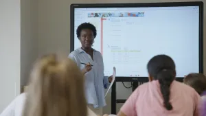 Een foto van presentator Tirsa die voor de klas staat. Achter haar staat een hele grote televisie die als een schoolbord gebruikt wordt.