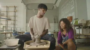 Een foto van presentator Serah die gehurkt zit naast Omar in een pottenbakstudio. Omar heeft voor zich een draaischijf in een plastic bak staan met daarop een met zijn handen gevormde vaas van klei. Omar heeft vlekken op zijn broek en armen van de klei.