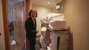 Een foto van presentator Pascal die op de gang staat van een hotel en naar een rolkast wijst waar allemaal opgevouwen schone was in en bovenop ligt.