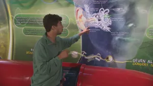 Een foto van Nizar bij een grote poster met daarop een menselijk gezicht waarvan het oor en de gehoorgang duidelijk zichtbaar zijn gemaakt. Ieder deel van de gehoorgang wordt op de poster beschreven. Nizar wijst naar het gedeelte dat "trommelvlies" heet.